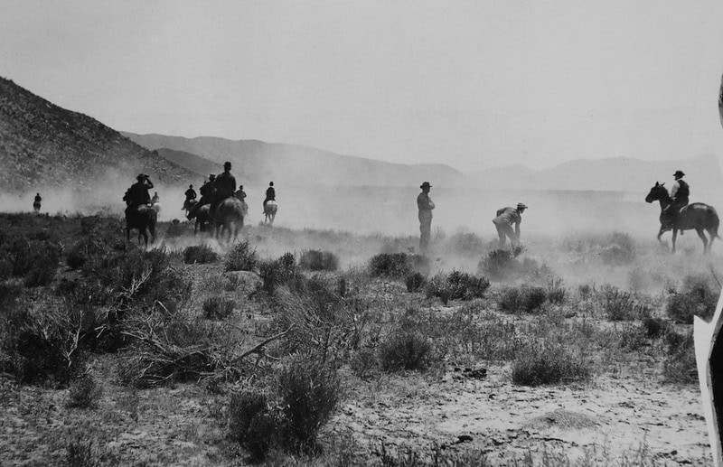 Horseback riders traversing a dusty desert landscape with mountains in the background.