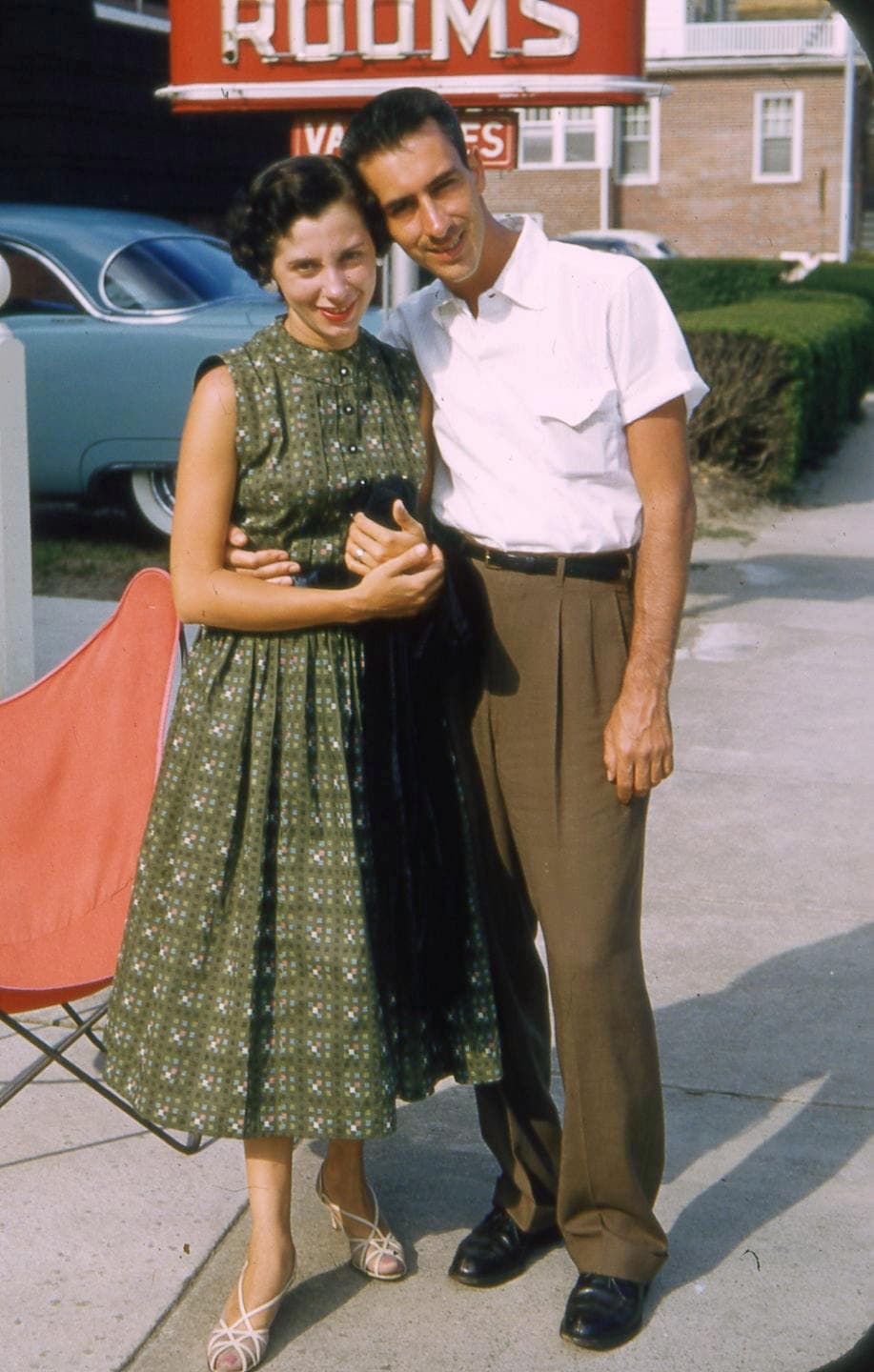Vintage couple standing outdoors, smiling, with a classic car in the background.