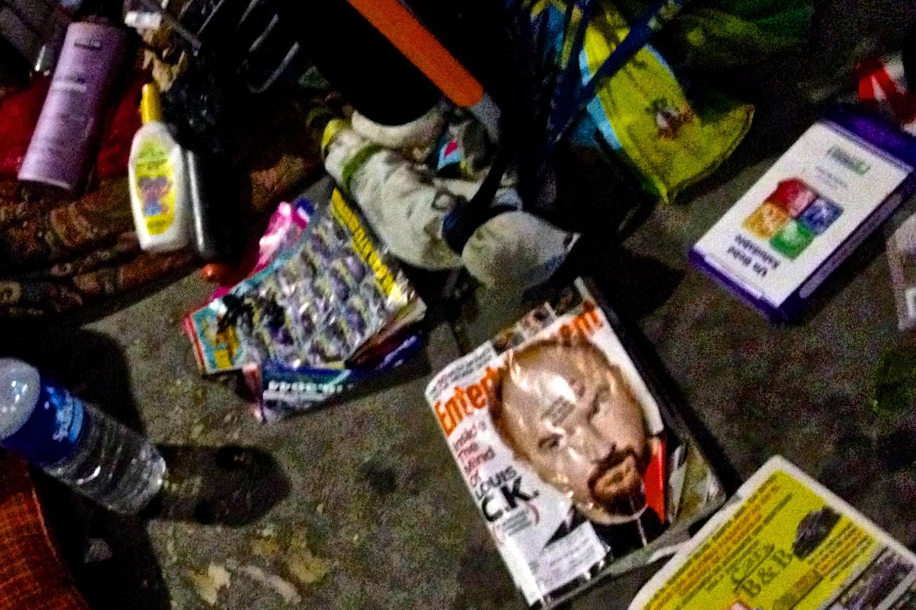 Cluttered shopping cart filled with groceries, magazines, and personal items in a busy store aisle.