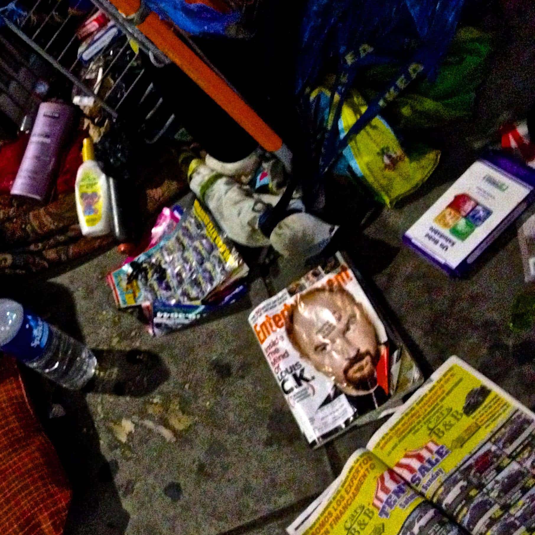 Cluttered shopping cart filled with groceries, magazines, and personal items in a busy store aisle.