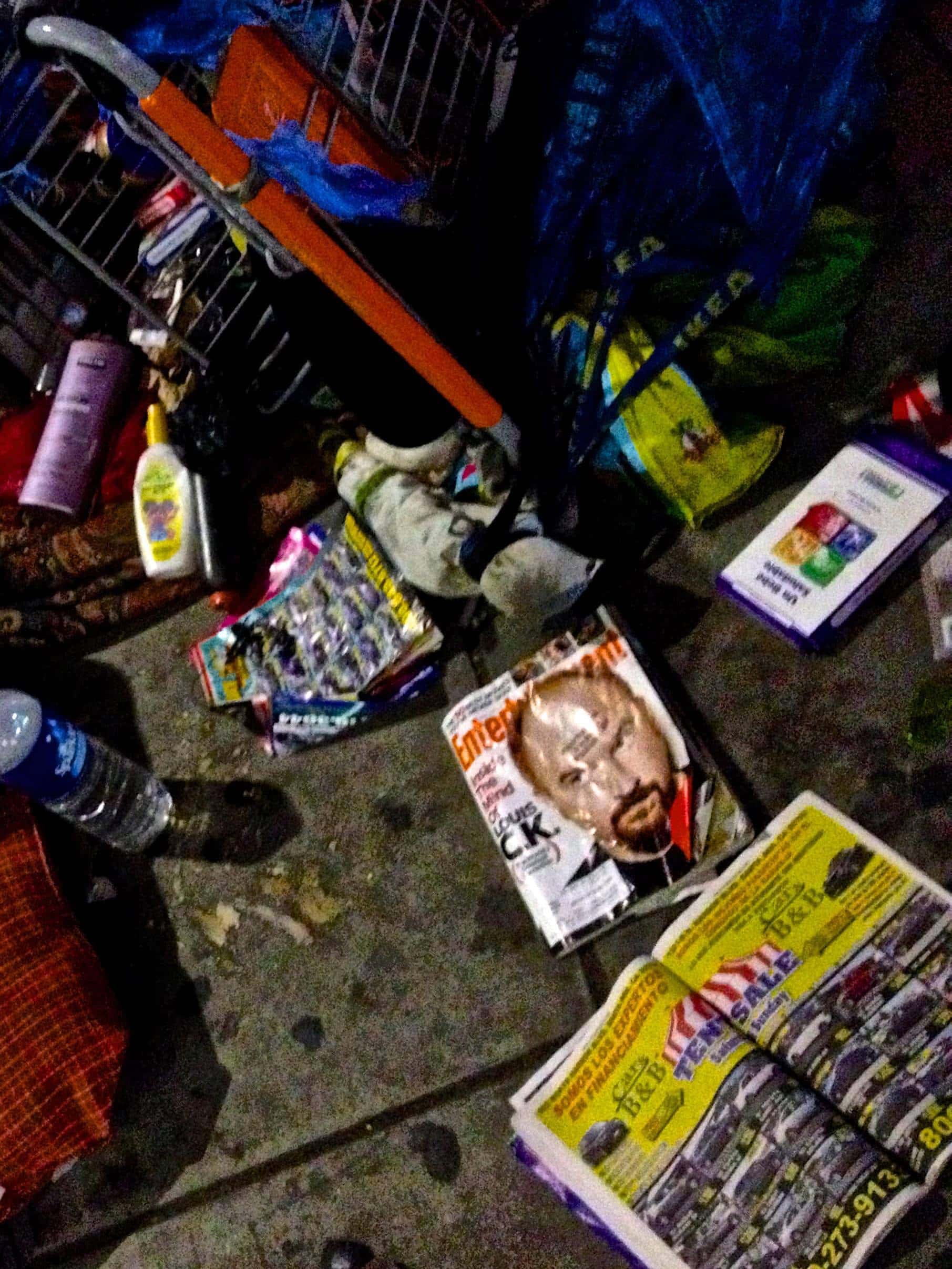 Cluttered shopping cart filled with groceries, magazines, and personal items in a busy store aisle.