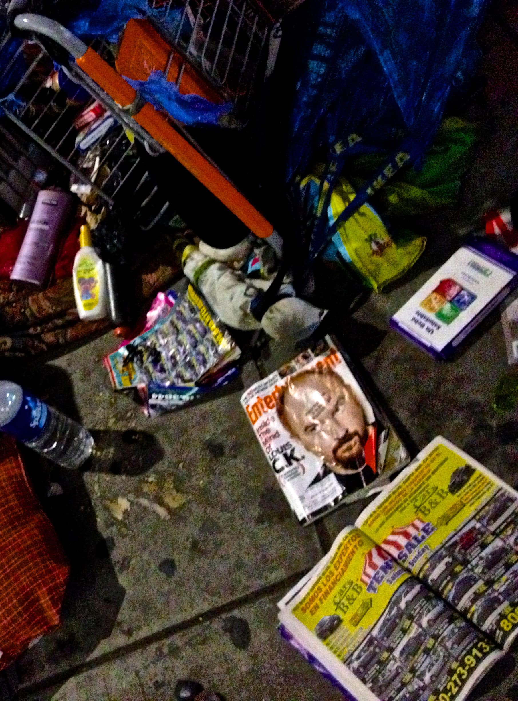 Cluttered shopping cart filled with groceries, magazines, and personal items in a busy store aisle.