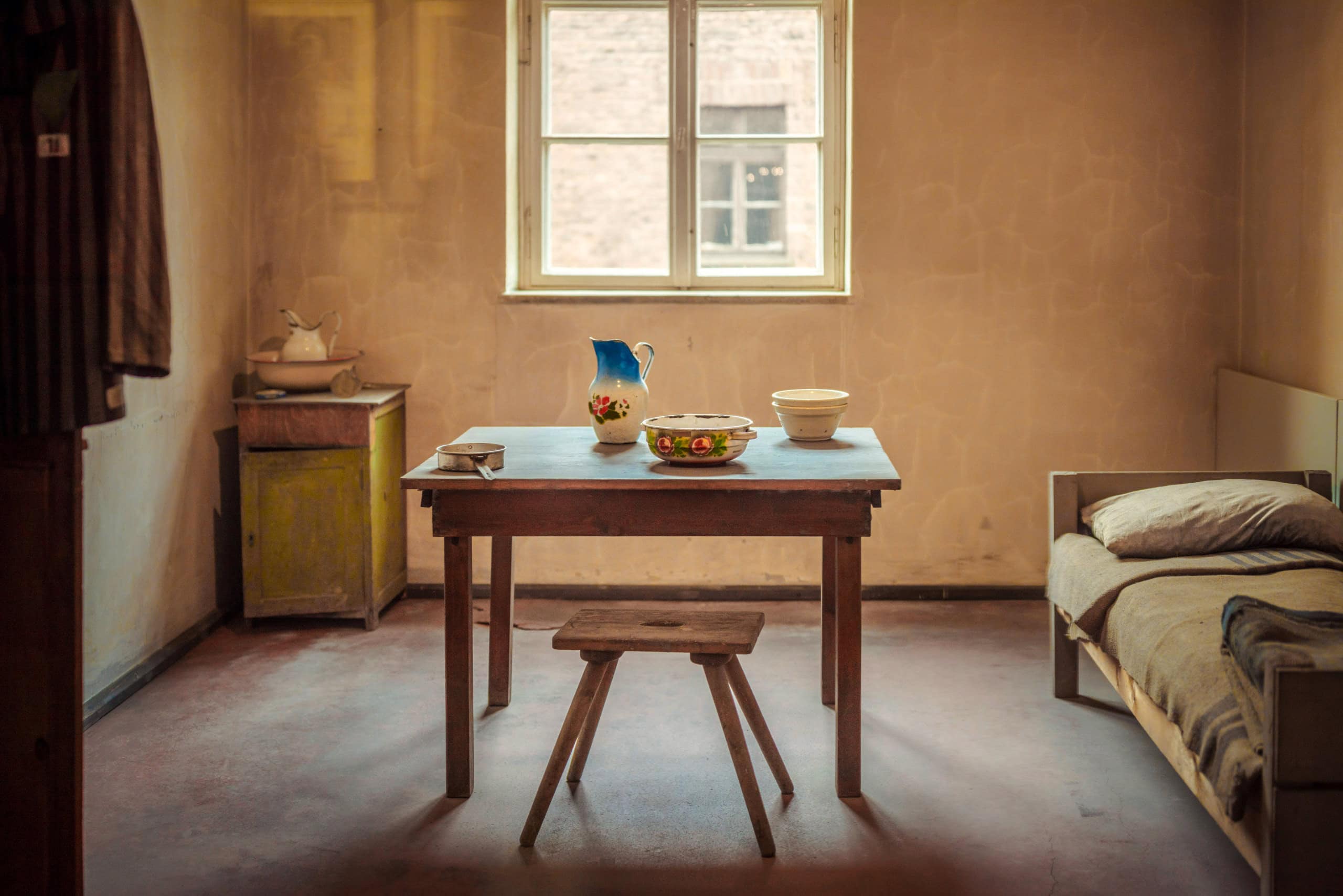 Minimalist rustic room with wooden table, stool, and bed near window.