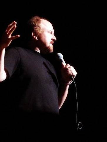 Louis C.K. holding a microphone, gesturing with his hand during a comedy routine.