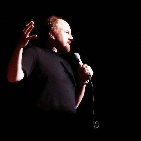 Louis C.K. holding a microphone, gesturing with his hand during a comedy routine.