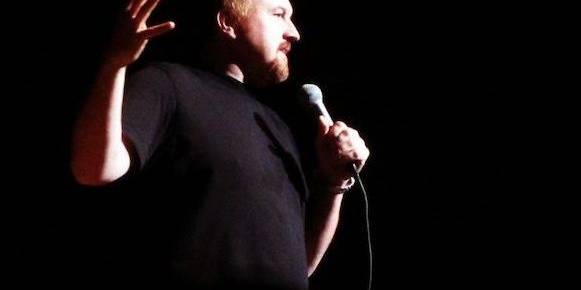 Louis C.K. holding a microphone, gesturing with his hand during a comedy routine.
