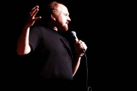 Louis C.K. holding a microphone, gesturing with his hand during a comedy routine.