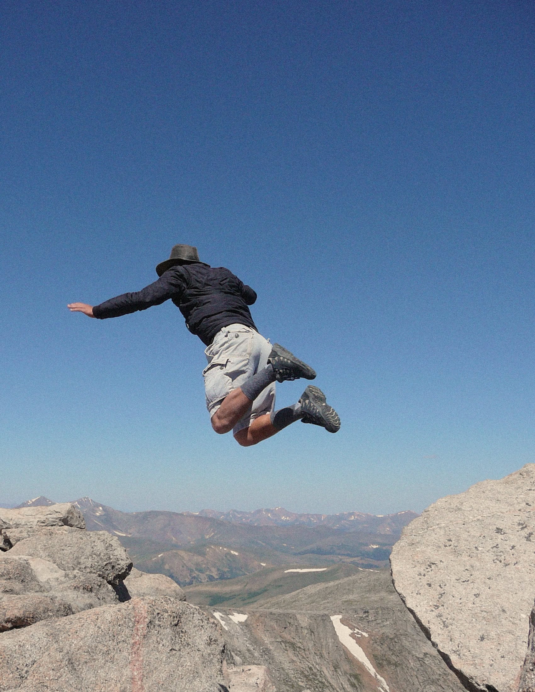 Person jumping midair over rocky mountain landscape under clear blue sky.